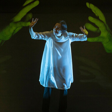 Chisato Minamimura in a white knee-length robe. To her left and right, she is bordered by two oversized green hands that are projected onto the dark wall behind her. She looks strained and, with her hands held up, gives the impression of wanting to push the projected hands away from her.