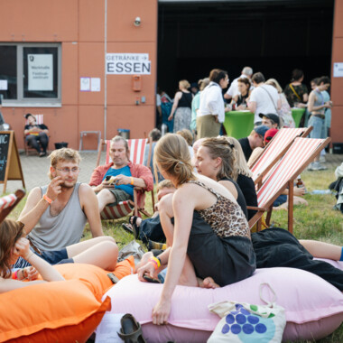 Several visitors are sitting on colourful beanbags and chatting on a lawn in front of a building. Other people are standing in front of the entrance, striking up conversations in small groups.