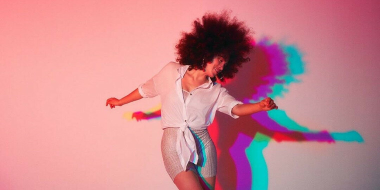 Selbstbeschreibung der abgebildeten Person: Eine schlanke Person of Colour mit einem großen braunen Afro tanzt. Der Hintergrund ist rosa und die Person trägt ein weißes Hemd und kurze Hosen. Die Person hat ein breites Lächeln im Gesicht und schaut nach links. Die Person sieht glücklich und energiegeladen aus.