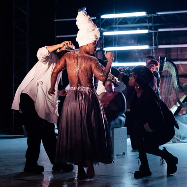 Bildbeschreibung seitens der Festival Theaterformen Redaktion. Selbstbeschreibung der abgebildeten Personen folgt. / Drei Schwarze Performerinnen auf der Bühne. Eine von ihnen wird von den zwei anderen mit einer braunen Flüssigkeit überschüttet. Sie ist oberkörperfrei, trägt einen langen Rock und steht mit dem Rücken zur Kamera. Im Hintergrund sind zwei weitere Performerinnen zu erkennen.