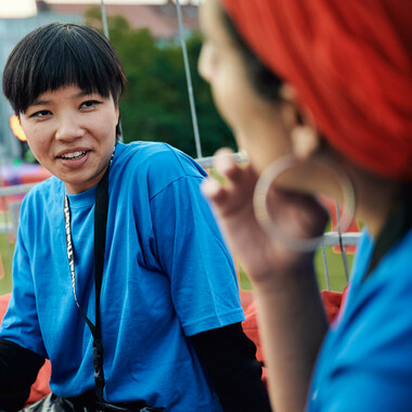 A person with short black hair in the left half of the photo. The person wears a blue tshirt and a black lanyard with the Festival Theaterformen logo on it. The person smiles.