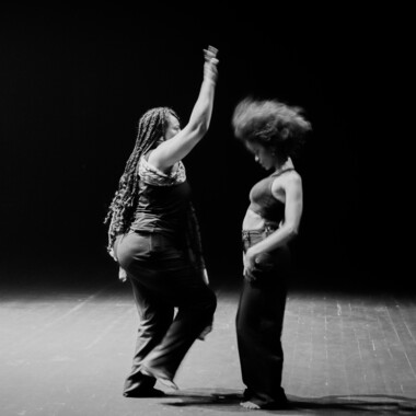 Ein Schwarzweiß-Foto aus dem Stück “When I saw the Sea” von Ali Chahrour: Es zeigt ein*e Performer*in in einem dunklen Raum an einem Marmortisch sitzen. Die Person trägt ein dunkles Tanktop, hält die Augen geschlossen und wirft den Kopf leicht nach hinten. Auf der Marmorplatte vor ihr ist die Reflektion der Person zu sehen.