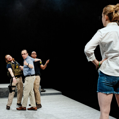 Bildbeschreibung seitens der Festival Theaterformen Redaktion. Selbstbeschreibung der abgebildeten Personen folgt. / Links im Hintergrund stehen drei Personen und schauen nach vorne. Zwei von ihnen tragen Kleidung mit der Aufschrift FBI. Auf der rechten Seite steht eine Person im Vordergrund und hat die Hände in die Hüften gestemmt. Sie trägt ein weißes Hemd und Jeansshorts und blickt in die Richtung der anderen drei Personen.