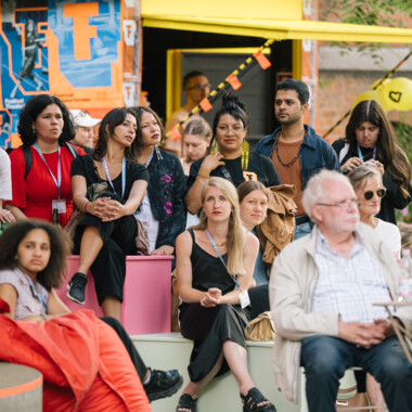 Eine Gruppe von Menschen sitzt aufmerksam auf farbigen Stufen im Freien während einer Veranstaltung. Im Hintergrund sind orange-blaue Festivalplakate sowie ein gelbes Vordach zu sehen. Einige Personen tragen Festivalausweise um den Hals. Die Stimmung wirkt konzentriert und interessiert.