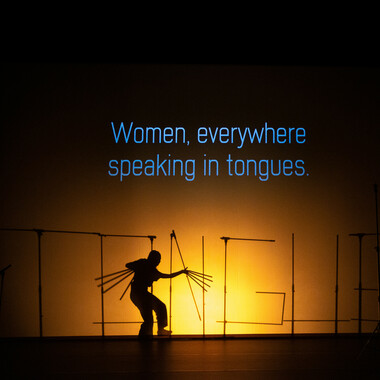 Image description by the editorial staff of Festival Theaterformen. Self-description of the depicted persons follows. / A theatre stage with a projection on a big screen of the phrase, "Women, everywhere speaking in tongues." in blue font. Underneath the projection the silhouettes of several microphone stands and a person can be seen. The microphone stands are arranged to form the word "TONGUE". In front of the microphone stands, there is a person in a bent posture with artificially extended fingers.