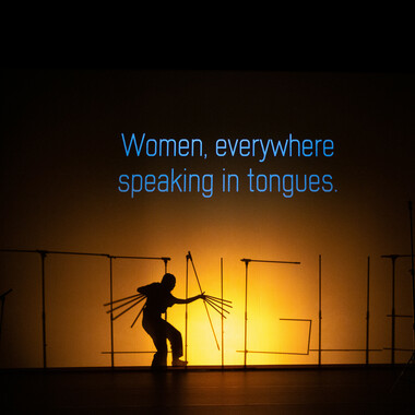 Bildbeschreibung seitens der Festival Theaterformen Redaktion. Selbstbeschreibung der abgebildeten Personen folgt. / Eine Theaterbühne mit einer Projektion des Satzes „Women, everywhere speaking in tongues.“ in blauer Schrift auf eine große Leinwand. Unterhalb der Projektion sind die Silhouetten mehrerer Mikrofonständer und einer Person zu sehen. Die Mikrofonständer sind so angeordnet, dass sie das Wort "TONGUE" bilden. Vor den Mikrofonständern befindet sich eine Person in gebeugter Haltung mit künstlich ausgestreckten Fingern.