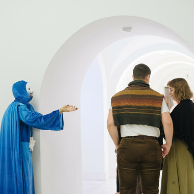 Ein weißer, heller Raum mit Bogengang, in dem drei Personen stehen. Links ist eine Person von der Seite zu sehen, die die Handinnenflächen zu einer bettelnden Geste zusammenführt und dabei das gegenüberstehende Paar anschaut. Sie trägt eine weiße Maske und ist von Kopf bis Fuß in ein blaues, fließendes Gewand gehüllt. Um den Hals der Person ist eine silberne Kette zu sehen, an der eine großer weißer Stein hängt.
