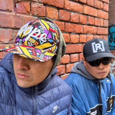Das Hiphop-Duo Eskina Qom: Sie sitzen vor einer Backsteinwand und tragen sportliche Kleidung. Die Person links blickt nach links und trägt eine bunte Cap sowie eine hoch zugeschlossene dunkelblaue Nike-Jacke. Die person daneben hat eine dunkle Sonnenbrille auf der Nase und trägt eine dunkle Cap, die mit dem Buchstaben R versehen ist. Die Person trägt eine blaue Jacke, darunter einen grauen Kapuzenpulli.