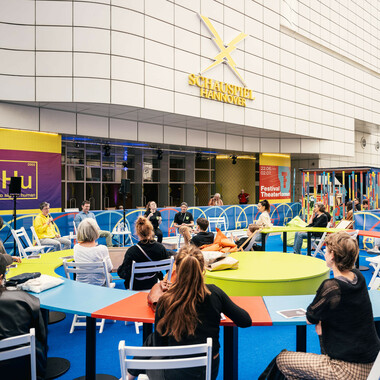A crowd of people at colourful, curved tables in the festival centre. The façade of the Schauspielhaus can be seen in the background.