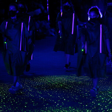 Several people in knee-length colorful capes with reflectors running vertically down the front. The people appear to be marching, they are wearing black masks. They hold green laser pointers in their hands that illuminate the ground beneath their feet. From behind they are illuminated with purple spotlights.