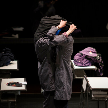 Two schoolgirls in dark school uniforms in the center of the picture. They stand closely together in the midst of white desks. They are exchanging their hijabs (head covers). The fabric of their hijabs fully covers the girls faces. A purple backpack rests on the desk behind them.