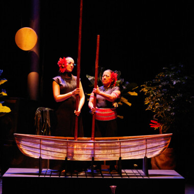 Zwei Personen stehen hinter einem hölzernen Boot auf einer Bühne. Beide halten lange Holzstangen und tragen ein dunkles Kostüm und eine auffällige große rote Blume im Haar. Im Hintergrund sind große Pflanzen und eine gelbe, runde Scheibe. Die Szene ist in warmes, stimmungsvolles Licht getaucht.