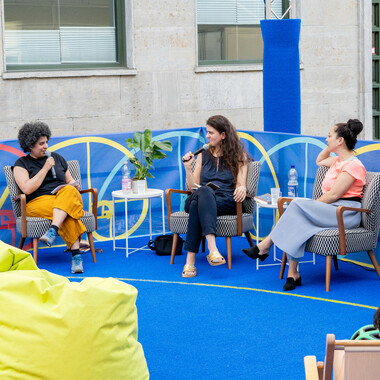 Von links nach rechts: Verena Meyer, Melanie Micudaj und Golschan Ahmad Haschemi. Sie sitzen im bunten Festivalzentrum auf der Prinzenstraße und führen den ersten Talk der Toolbox-Gesprächsreihe, Golschan spricht gerade in ein Mikrofon. Vor ihnen sind grüne und orange Sitzsäcke sowie eine grüne runde Bassplattform zu sehen. Das Festivalzentrum ist blau.