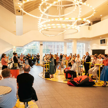 Viele Menschen im Schauspielhausfoyer. Über ihnen hängt ein großer geschwungener und abstrakter Kronleuchter, der hell erleuchtet ist. Rita Mazza steht links im Bild und gebärdet DGS.