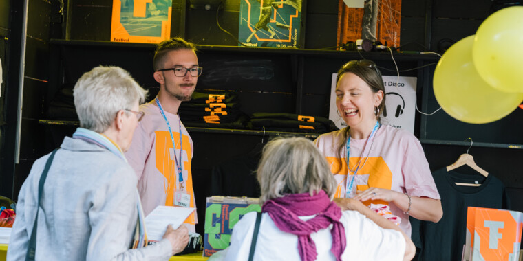 Infobox of the Festival Theaterformen. Four people, two in the box, two in front of the box are acting together. One person is laughing. There are posters in the background, folded T-shirts and program booklets on the side and balloons hanging in the corner on the right.