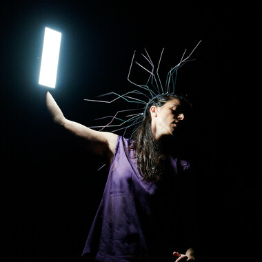 Bildbeschreibung seitens der Festival Theaterformen Redaktion. Selbstbeschreibung der abgebildeten Personen folgt. / Eine Person, die sich mit einer sehr grellen Tageslichtlampe selbst anleuchtet. Der Hintergrund ist komplett dunkel. Die Person hat lange, dunkle Haare, trägt ein lila Tanktop und eine Krone aus langen, verzweigten Drähten auf dem Kopf. Sie hält die Lampe mit ihrer rechten Hand, beleuchtet sich selbst damit von der Seite und hält ihren Kopf in die entgegengesetzte Richtung. Ihre Augen sind geschlossen.