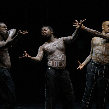 Three PoC side by side, their naked torsos covered with white lettering. They wear black pants and perform expressive, indefinable gestures and mimics with their arms and faces. At the same time, their fingers are tense.