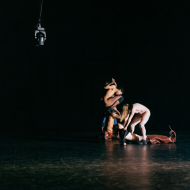 Mehrere nackte Performer bilden eine dichte, ineinander verschlungene Gruppe am Boden einer dunklen Bühne. Körper sind übereinander geschichtet und in einer skulpturalen Pose miteinander verbunden. Rechts im Bild hängt ein Scheinwerfer von der Decke herab.