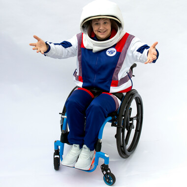 Bildbeschreibung seitens der Festival Theaterformen Redaktion. Selbstbeschreibung der abgebildeten Personen folgt. / Eine weiße Person im Rollstuhl mit ausgestreckten, offenen Armen. Sie trägt ein Astronautenanzug.