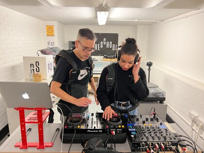 DJ-Workshop mit Troi Lee: Links ist der DJ Troi Lee mit einer am Workshop teilnehmenden Person (rechts) zu sehen. Beide tragen Woojer Westen und stehen hinter einem Tisch auf dem diverses DJ-Equipment und ein Laptop liegt. Die Person rechts bedient einen DJ-Mixer.