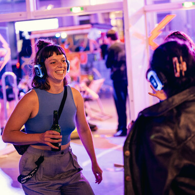 A person with blue glowing headphones. She smiles and seems to be dancing. She is holding a green bottle in her hand.