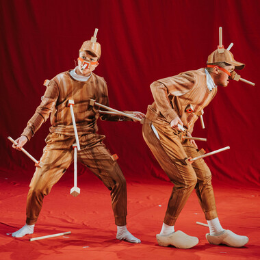 Zwei Performer*innen: Rosana Cade (they/them) und Ivor MacAskill (he/him) stehen auf einer Bühne, die mit leuchtend rotem Stoff bespannt ist. Rosana (links) ist eine weiße nicht-binäre Person mit kurzen Haaren und Ivor (rechts) ist ein weißer Mann mit kurzen Haaren und roten Bartstoppeln. Beide tragen Trainingsanzüge mit Holzmaserung und Caps, aus denen echten Holzstöcke herausragen. Rosana steht uns gegenüber und hat einen fragenden Gesichtsausdruck. Ein hölzerner Stock, der aus dem Holzwürfel auf Rosanas Nippel herausragt, ist mit dem Holzwürfel an Ivors Po verbunden. Ivor steht seitlich, von Rosana abgewandt, und scheint mitten im Gehen erstarrt zu sein.