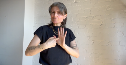 Rita Mazza in front of a white stone wall. Rita wears a black T-shirt and looks into the camera. With her hands, Rita makes a gesture in DGS.