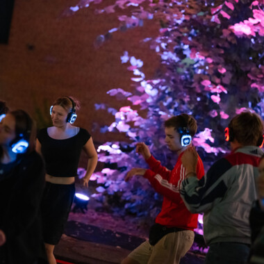 Mehrere Personen tanzen mit leuchtenden Kopfhörern bei einer nächtlichen Silent Disco im Freien. Im Hintergrund ist ein farbig angestrahlter Baum zu sehen, der die Szene in violettes Licht taucht.