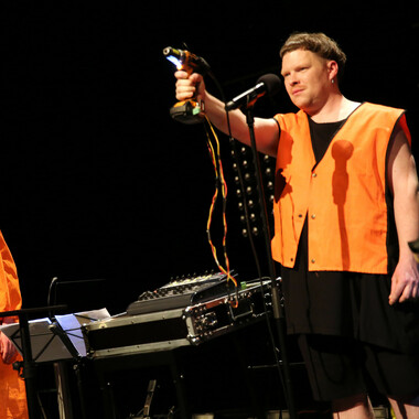 Image description by the editorial staff of Festival Theaterformen. Self-description of the depicted persons follows. / On the right there is a person wearing black clothes and a neon orange vest. They are holding a drill in their right hand. The drill is connected to a sound desk via two visible and colorful cables. On the left there is a music stand with white sheets on it.