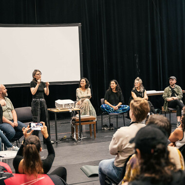 Rita Mazza steht in einem Halbkreis inmitten der weiteren Künstler*innen, die am Sign Language Art Laboratory geforscht haben. Rita gebärdet, vor Rita ist ein sitzendes Publikum zu sehen. Alle schauen zu Rita.