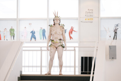 Eine Person steht auf eine der Treppen im Foyer des Schauspielhauses. Ihr gesamter Körper ist weiß geschminkt, sie trägt einen Kopfschmuck, der an eine Krone erinnert. 