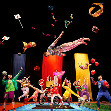 Many colorfully dressed performers on a stage. On multicolored patterned carpets, they perform various acrobatic movements and appear to be very cheerful. Above their heads, various objects such as shoes, road signs, balls and clothes are floating wildly. One performer is in the air and about to do a somersault. In the background of the picture, DJ Dino can be seen in front of a mixing desk.