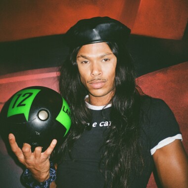 Ein Mann mit langen Haaren und einem schwarzen Barett hält einen schwarzen Bowlingball mit grüner Aufschrift „12“ in der Hand. Er trägt ein enges schwarzes T-Shirt mit weißem Kragen und einem Schriftzug auf der Brust. Der Hintergrund ist in rotes Licht getaucht, was der Szene einen dramatischen Effekt verleiht.