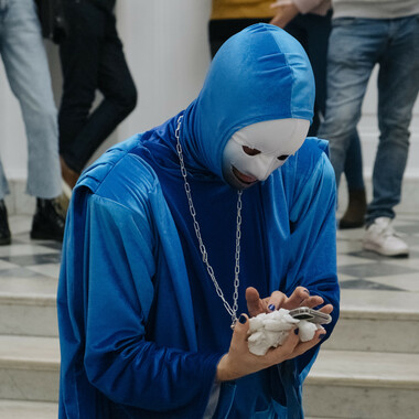 Eine kostümierte Person auf einer Marmortreppe, dessen Gesicht von einer weißen Maske verdeckt wird. Die Person trägt ein fließendes Gewand in der Farbe Blau und tippt auf ihr Handy, das in ihrer rechten Hand liegt und an eine silberne Kette um seinen Hals hängt. Im Bildhintergrund sind die Beine anderer Menschen zu sehen.