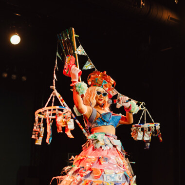 Eine Person auf einer Bühne. Sie trägt ein auffälliges Kostüm aus bunten Materialien, es besteht aus einem ausladenden Reifrock, einem blauen BH und einer Konstruktion aus zwei große, dekorative Objekte zwischen die Wimpelgirlanden gespannt wurden. In der eine Hand hält sie ein Mikrofon, in der anderen eine Wasserspritzpistole. 