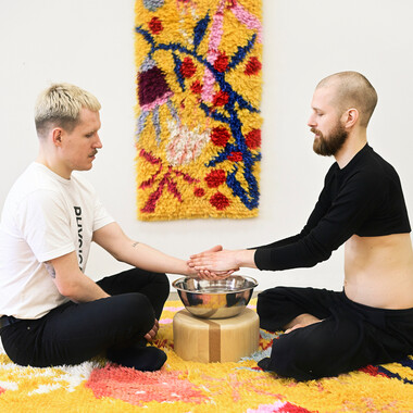 Self-description of the depicted persons: Two white, able-bodied cis men are sitting cross-legged on a colorful carpet. There is a metal bowl on a wooden pedestal between them and a colorful tapestry in the background. The man on the right is washing the left man's left hand. 
