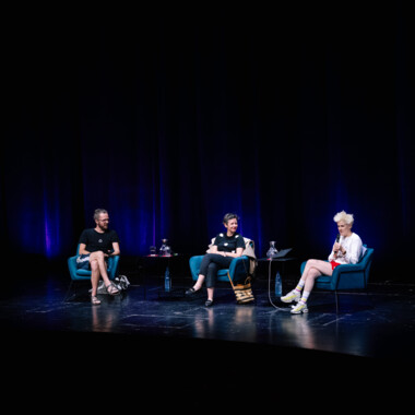 Three people sit in chairs on a dark stage, engaged in a conversation.