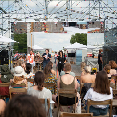 Students from the Festivalcampus Niedersachsen during a discussion session at the Theaterformen festival. They are sitting outside on wooden chairs; three people are standing at the front, one of whom is speaking into a microphone.