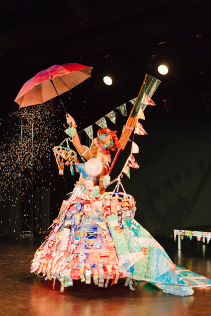 Person steht auf einer Bühne. Sie trägt ein buntes, aus recycelten Materialien gefertigten Kostüm mit großem Rock und vielen dekorative Objekte an denen viele Wimpelgirlanden befestigt wurden. In einer Hand hält sie einen roten Regenschirm. 