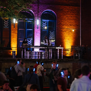 Menschen mit leuchtenden Kopfhörern tanzen bei einer nächtlichen Silent Disco im Hof eines Gebäudes mit orange-violetter Beleuchtung. Im Hintergrund stehen zwei DJs auf einem Balkon vor großen Fenstern und legen Musik auf.
