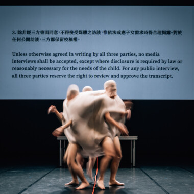 Mehrere Personen stecken zusammen in einem hautfarbenen, sackähnlichen Kostüm, durch das nur ihre Hände und Beine sichtbar sind. Durch die Bewegung sind sie leicht verschwommen Eine Projektion im Hintergrund zeigt englischen und chinesischen Text.