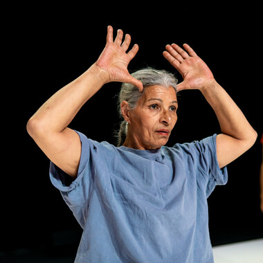 The dancer Mimouna Latifa Khamessi in a blue, loose tshirt. Her gray hair is tied back into a low ponytail, she her hands up: her thumbs rest at her temples, her palms face outward. She gazes into the distance.