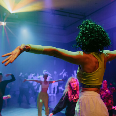 A club room immersed in colorful party lights. In the foreground, dancing performer Anna Seymour can be seen from behind. She is wearing a sleeveless and cropped training top and white track pants. Her shoulder-length hair is curly and black. She stretches her arms out wide. In the background of the picture, a large number of people can be seen who are imitating Anna's movements.