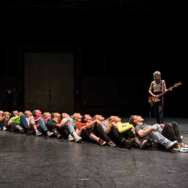 Stage situation: A large number of teenagers sit one behind the other on the floor, each in the lap of the person sitting behind them. Next to them is a person with an electric guitar.