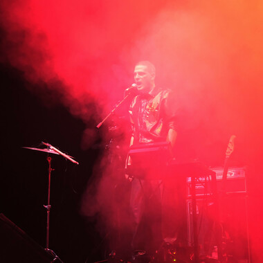 A concert situation. Alen Sinkauz on stage, singing into a microphone, Alen’s mouth is open. He has very short hair and is a dark leather costume. The stage is covered in fog.