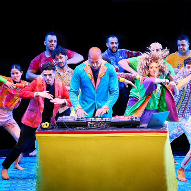 DJ DINO performs in an eye-catching blue party costume. In front of him there is a mixer on a table covered with yellow fabric. Next to it a laptop and headphones can be seen. Behind him, there are colorfully dressed performers. They are dancing barefoot. With their arms bent and palms touching in front of the chest, they all perform the same movement.