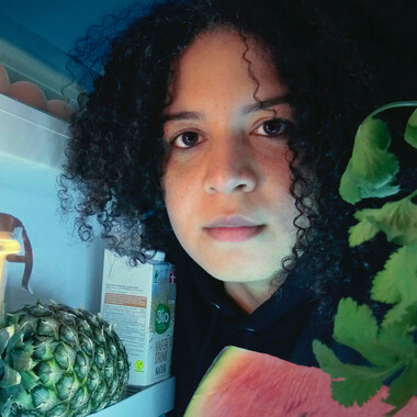 Nahaufnahme der Performerin aus einem Kühlschrank heraus. Sie blickt in den Kühlschrank und direkt in die Kamera. Ihr Kopf ist umgeben von Lebensmitteln: Kräuter, Hafer Drink, Ananas, Wassermelone.