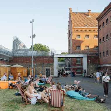Besuchende sitzen und liegen auf Liegestühlen und Sitzsäcken auf einer Wiese im Festivalzentrum in der JVA Rennelberg. Im Hintergrund sind Backsteingebäude und Stacheldraht.