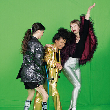 Auf dem Bild sind drei Frauen: Alice Giuliani, Victoria Antonova und Laila White (von links nach rechts), die vor einem neongrünen Hintergrund stehen. Alice Giuliani ist mit dem Rücken zur Kamera gedreht. Victoria Antonova trägt einen goldenen Anzug und stützt sich auf eine Krücke und lacht dabei. Alice Giulianis Hand liegt auf ihrer Schulter. Ganz rechts ist Laila White die eine Tanzbewegung Richtung Victoria Antonova macht und einen Umhang mit Federn und eine silberfarbene Hose trägt.