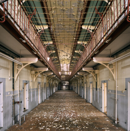 An interior view of Rennelberg Prison. A long, symmetrical prison corridor with cell doors on both sides, and peeling paint on the floor.